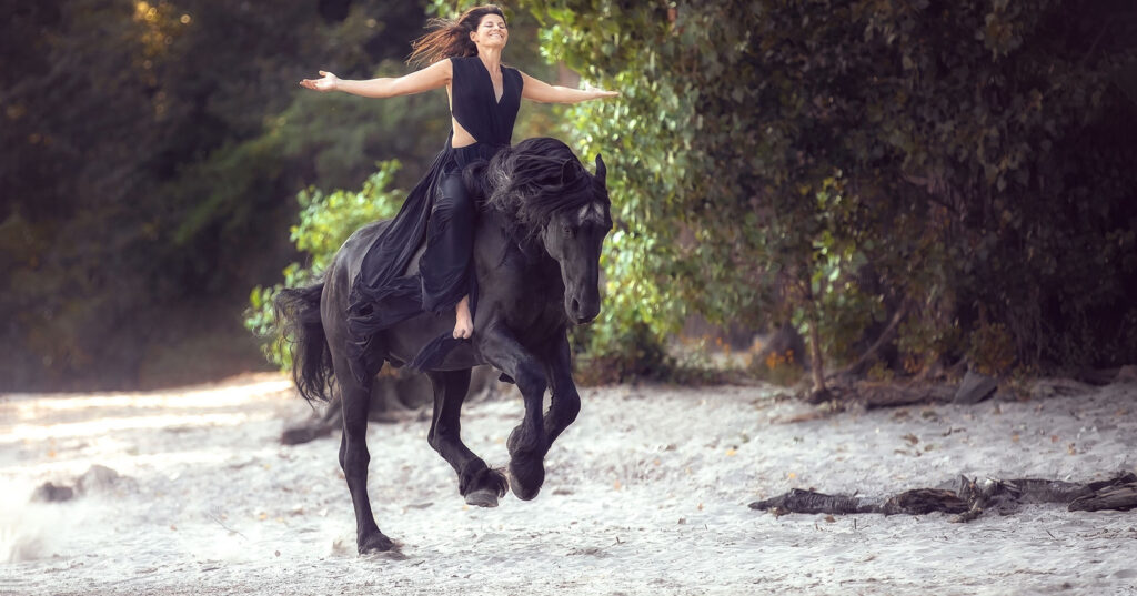 Alex und Macho auf Sand Eine Frau reitet auf einem Friesen ohne Sattel und Trense an einem Strand.
